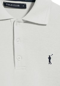 Vit polo skjorta med texturerad tyg, treknappsslåk och en liten marinblå broderad figur inom designen. "POLO CLUB"-etikett synlig.