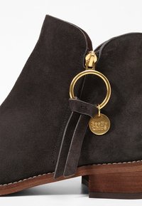 Mörkgrå mockaskor vid vristen med en sidodragkedja, träklack och en guldfärgad hårdvaruring med en etikett som säger "See By Chloé."