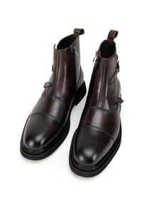 Botas de tobillo de cuero marrón con un panel trasero texturizado, puntera afilada, hebillas decorativas y una suela gruesa de goma negra.
