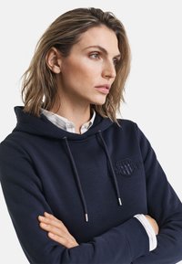 Marineblå hettegenser laget av mykt stoff, med et brodert logo, frontsnører med sølvdetaljer, og en lagdelt hvit skjorte under.