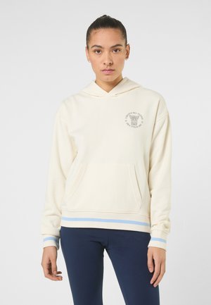 Jonge vrouw die een crèmekleurige hoodie met voorzak en blauw gestreepte manchetten draagt, gecombineerd met donkerblauwe broek, tegen een effen achtergrond.