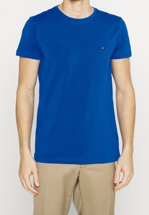 Mężczyzna ubrany w jednolity, niebieski T-shirt z krótkim rękawem z małym logo na piersi oraz beżowe spodnie, na białym tle.