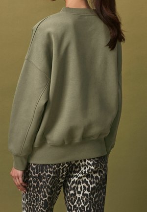Mujer con cabello oscuro recogido en una coleta, vistiendo una sudadera grande de color verde oliva y pantalones con estampado de leopardo, frente a un fondo en tono verde oliva a juego.