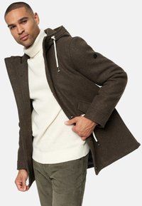 Jachetă dintr-un amestec de lână maro cu glugă și șnur, având o închidere cu fermoar, buzunare laterale și un pulover alb din tricot cu model cu nervuri dedesubt.