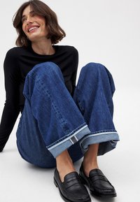 Blå denim vida jeans med uppvikta fållar, i kombination med svarta läderloafers. Modellen har på sig en figurnära svart tröja med lång ärm.