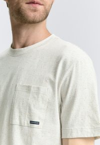 Tricou gri deschis cu dungi, cu gât rotund, având un buzunar pe piept și o etichetă mică cu logo. Material moale cu un model texturat, puncțat.