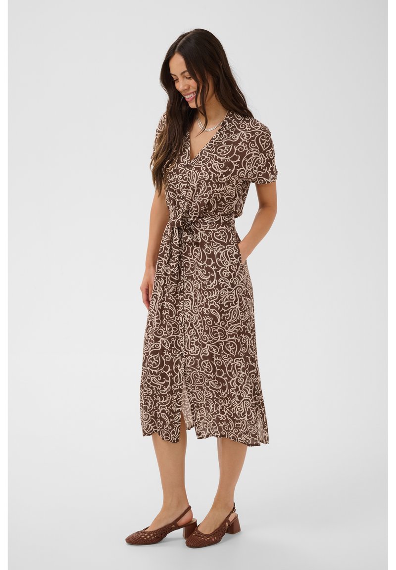 Femme souriante, portant une robe midi à motifs marron et beige avec manches courtes, ceinture à nouer devant, et des chaussures marron à talons carrés.