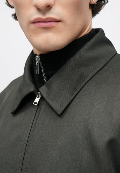 Sivá bunda so štruktúrovaným golierom, zapínaním na zips a rebrovaným čiernym vnútorným golierom. Hladký povrch s jemnými, nenápadnými vzormi.