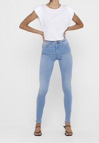 Vaqueros ajustados de color azul claro con cintura alta, confeccionados en denim. Combinados con una camiseta blanca de corte holgado y sandalias negras de tiras.