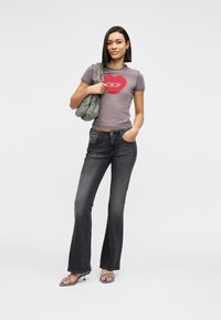 Νεαρή γυναίκα στέκεται με το ένα χέρι στην τσέπη και το άλλο κρατά μια τσάντα ώμου από τζιν, φοράει ένα διακοσμημένο γκρι μπλουζάκι και μαύρο τζιν.