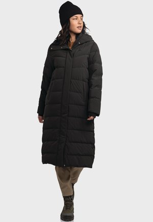 Langer schwarzer Puffer-Mantel aus gestepptem Material, mit hohem Kragen, Druckknöpfen vorne und lockerer Passform, kombiniert mit khakifarbenen Hosen und Stiefeln.