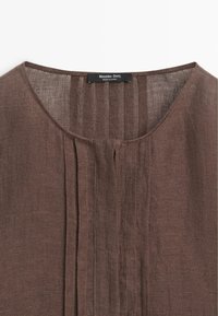 Bruine linnen blouse met ronde hals en verticale plooien in het midden voor, met een zwart "Massimo Dutti"-label aan de binnenkant van de kraag.