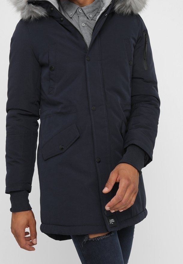 Man som bär en marinblå vinterjacka med huva som har pälsdetalj, med knappar och dragkedja samt framfickor, över blå jeans.