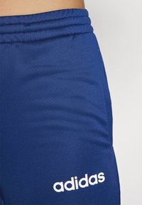 Jūras zilas sporta bikses ar teksturētu stiepļu audumu. Ar sakumpētu jostasvietu un baltu "adidas" logotipu apakšējā kājas daļā.