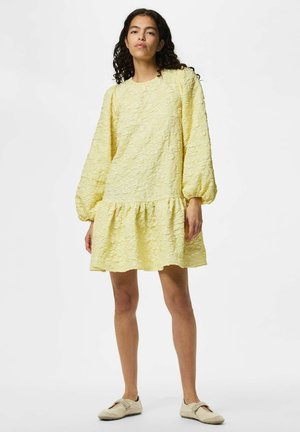 Jeune femme debout portant une robe mini jaune pâle texturée à manches longues bouffantes et des chaussures crème sans lacets.