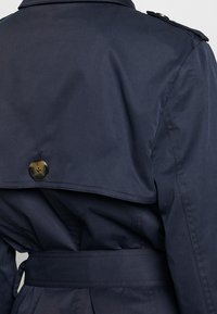 Tylny widok granatowego płaszcza trench z zapiętym klapą ochronną i pasującym materiałowym paskiem w talii.