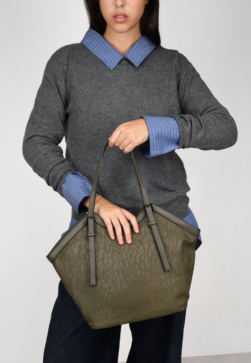 Bolsa de tote verde oliva con superficie texturizada y dos asas resistentes, sostenida por una persona que lleva un suéter gris con un collar a rayas azules.