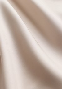 Tissu satin beige à la texture lisse ; présente un éclat doux et un tombé subtil, mettant en valeur des plis délicats et des reflets de lumière.