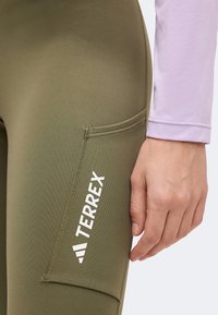 Gros plan d’une main reposant à côté d’un legging vert olive avec une poche latérale et le logo blanc « TERREX ».