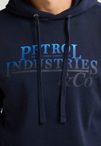 Hanorac cu glugă de culoare bleumarin, realizat dintr-un material texturat. Include un imprimeu grafic în nuanțe de albastru și gri, ce afișează textul "PETROL INDUSTRIES & Co™" și are șnururi ajustabile.