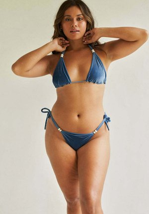 Kobieta poprawiająca włosy, ubrana w niebieski dwuczęściowy bikini z wiązanymi ramiączkami i ozdobami z koralików na gładkim, neutralnym tle.