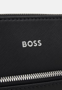 Černá syntetická peněženka s hladkým povrchem, stříbrným zipem a embosovaným logem "BOSS" na povrchu.