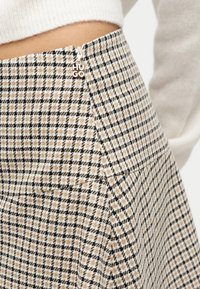 Κοντινό πλάνο σε φούστα με σχέδιο ψαροκόκαλο σε μπεζ και μαύρο, με πλαϊνό φερμουάρ και λογότυπο "HUGO", φορεμένη με πλεκτό πουλόβερ σε εκρού απόχρωση.