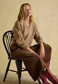 Brązowy, strukturalny sweter z dekoltem w kształcie litery V w połączeniu z brązowymi, luźnymi spodniami z szerokimi nogawkami oraz różowymi butami w wzory, siedzący na czarnej, drewnianej krześle.