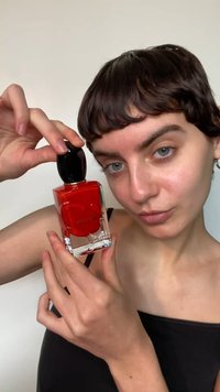 Flacon de parfum rouge avec un bouchon noir tenu par une personne. Forme carrée, base en verre transparent, avec un logo sur le devant.