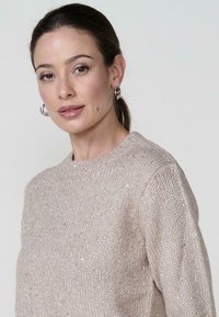 Suéter de punto beige con cuello redondo, que presenta sutiles destellos dorados en toda la tela y puños acanalados para aportar textura.