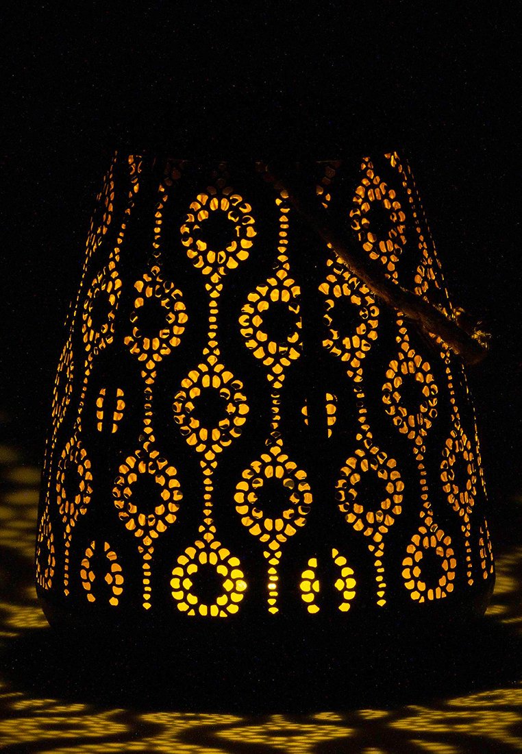 Czarna metalowa latarnia z misternymi wzorami wyciętymi, emitująca ciepły pomarańczowy blask z wewnętrznego źródła światła, tworząca szczegółowe cienie.