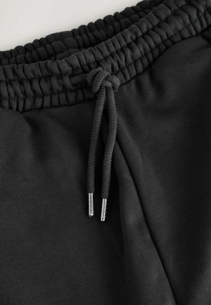 Zwarte sweatpants hebben een verzamelde elastische tailleband met een geweven koord, eindigend in zilveren uiteinden. De stof lijkt zacht en glad.