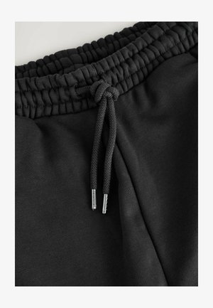 I pantaloni della tuta neri presentano una vita elastica arricciata con un laccetto intrecciato, terminante con punte argentate. Il tessuto appare morbido e liscio.