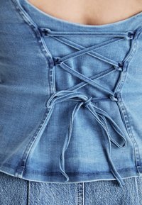 Lichtblauwe denim top met een kantontwerp aan de voorkant. De stof is zacht, met een textuur, en heeft gekruiste veters en een ronde halslijn.