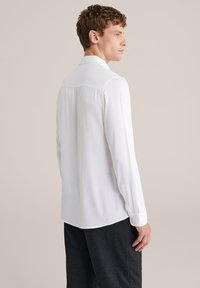 Camicia bianca a maniche lunghe con yoke posteriore e pieghe sottili, tessuto testurizzato, polsini con bottoni e orlo curvo, abbinata a pantaloni scuri.