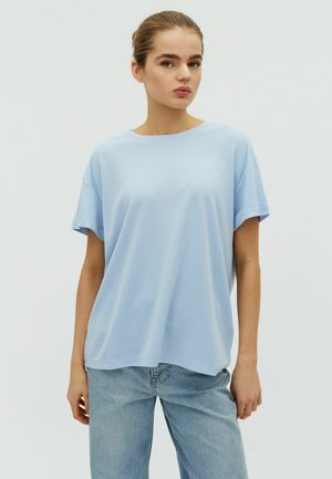 Junge Frau trägt hellblaues T-Shirt mit kurzen Ärmeln und lockere blaue Jeans, steht vor einem einfarbigen hellen Hintergrund.