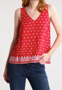 Haut rouge sans manches avec un col en V, présentant un motif géométrique blanc et des accents floraux à l'ourlet. Porté avec un jean bleu.