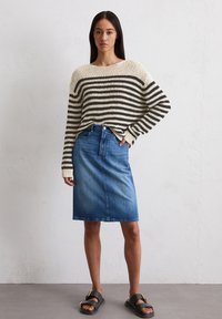Gestreepte gebreide trui in crème en bruin, gecombineerd met een denim kokerrok. Sandalen hebben een zwarte band en een metallic accent.