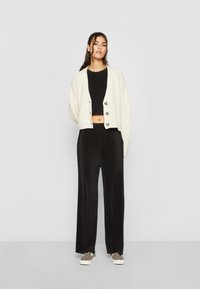 Krémový pletený kardigán s ryhovanou textúrou, čierny crop top, čierne široké nohavice a kockované slip-on topánky. Jednoduché gombíky na kardigáne.