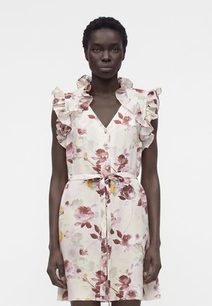 Γυναίκα με αμάνικο, φλοράλ φόρεμα με βολάν στους ώμους και ζώνη στη μέση, στέκεται μπροστά σε ένα απλό λευκό φόντο.