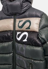 Dunkle Daunenjacke mit beigen und schwarzen Streifen, großen "S"-Buchstaben auf der Brust, Frontreißverschluss und hohem gepolstertem Kragen.