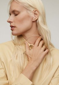 Camisa de cuero amarillo con un cuello alto, adornada con botones, combinada con una mano que muestra un delicado anillo. Cabello rubio largo y liso.