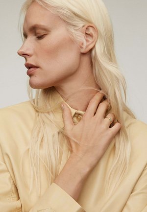 Camisa de cuero amarillo con un cuello alto, adornada con botones, combinada con una mano que muestra un delicado anillo. Cabello rubio largo y liso.