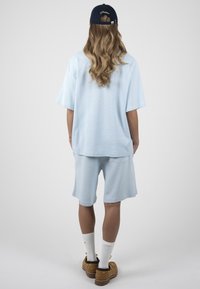 Personne aux longs cheveux ondulés portant une casquette bleu marine, un t-shirt bleu clair oversize et un short, des chaussettes blanches et des chaussures marron, debout de dos.