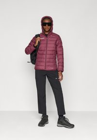 Μαρούλι μπουφάν με κουκούλα, με φερμουάρ και λεπτομέρεια λογότυπου; σε συνδυασμό με μαύρο παντελόνι και στιβαρές μαύρες μπότες πεζοπορίας.