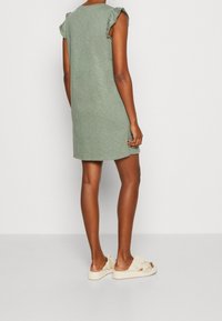 Robe verte longueur genou avec manches courtes à volants, en coton texturé. Portée avec des chaussures légères sans lacets de couleur claire.