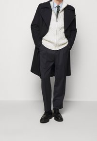 Mörk ullkappa över en vit kabelstickad tröja, i kombination med marinblå klädkostymbyxor och svarta läderskor. Enkla, moderna design detaljer.