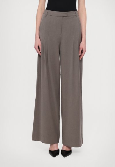 Pantaloni cu pantaloni largi dintr-un material bej deschis, cu pliseuri frontale, un talie structurat și o textură netedă. Purtați cu tocuri negre.