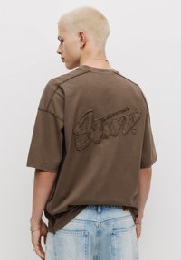 Hnědá bavlněná oversized tričko s texturovaným vyšitým logem na zadní straně. Krátké rukávy a uvolněný střih, spojené s modrými džínami.