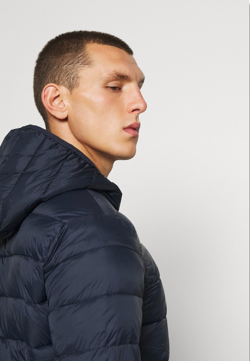 Veste matelassée navy avec capuche, présentant une texture lisse et des détails de couture horizontale. Modèle présenté de profil.